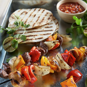 Grilled meat and vegetable kabobs on skewers on a tray with grilled soft tortillas and salsa