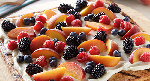 Square pizza on grilled crust. Topping is a white creamy base with peaches and berries arranged on top