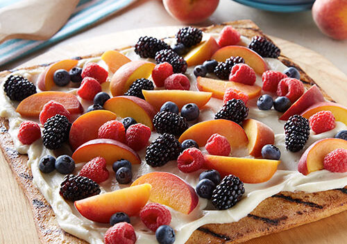 Square pizza on grilled crust. Topping is a white creamy base with peaches and berries arranged on top