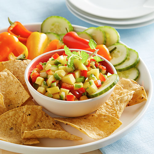 Mango salsa in a bowl surrounded by taco chips, sliced and seeded chili peppers and cucumber slices